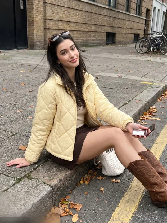 Girl in butter yellow quilted jacket sitting on city curb with leather boots and woven tote, autumn city sidewalk ChatGPT photo prompt
