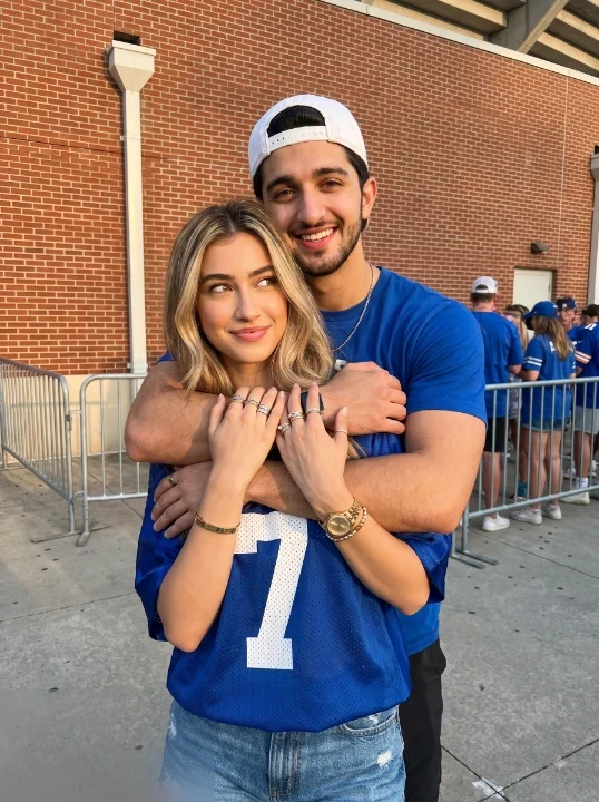 ChatGPT couple photo prompt man and woman in matching royal blue jersey and backwards cap hugging outside brick stadium wall