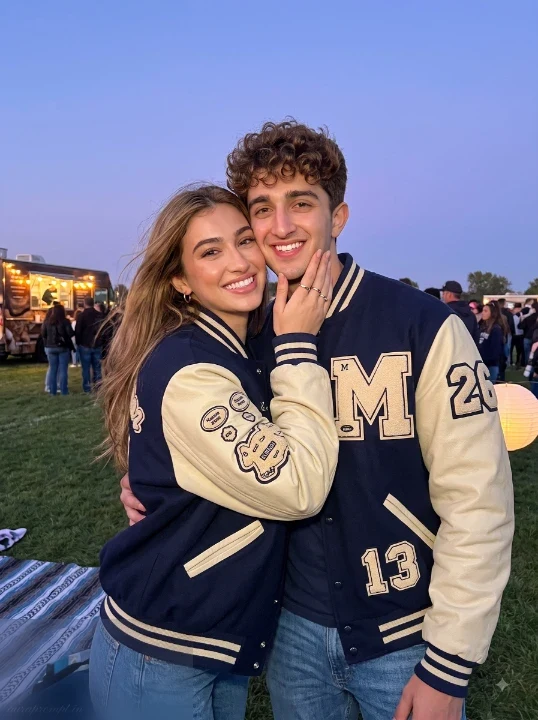 AI tailgate party photo prompt couple in matching navy letterman jackets on grassy field at dusk with lantern light background